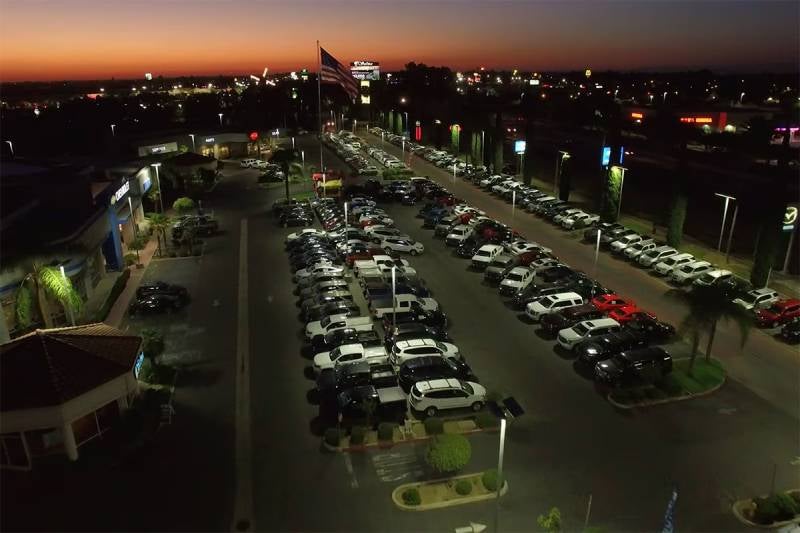 Aerial nighttime view of a car dealership lot filled with parked vehicles, with streetlights and a sunset in the background