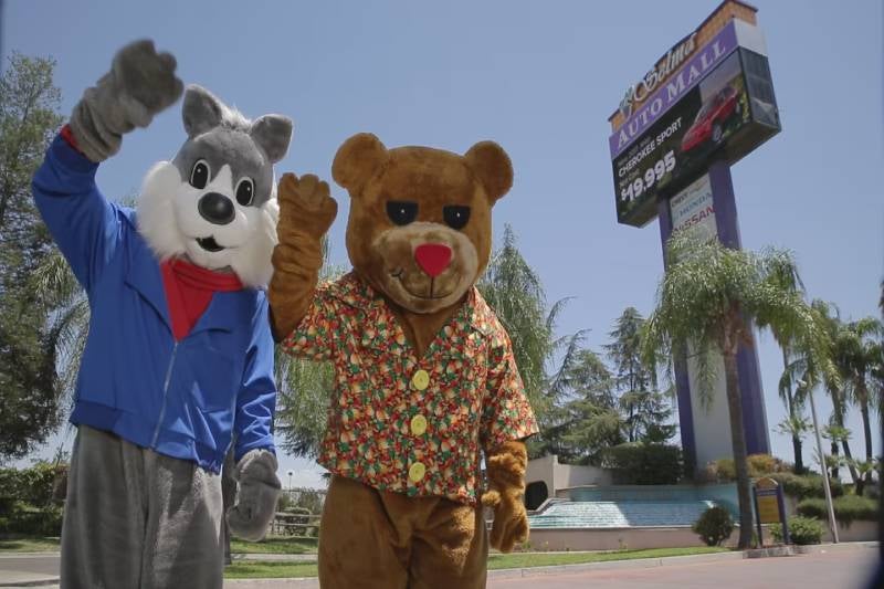 Mascot characters waving in front of an auto mall sign.