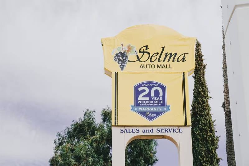 Outdoor sign for Selma Auto Mall featuring a yellow banner with grapes, a 20-year/200,000-mile warranty badge, and ‘Sales and Service’ written below