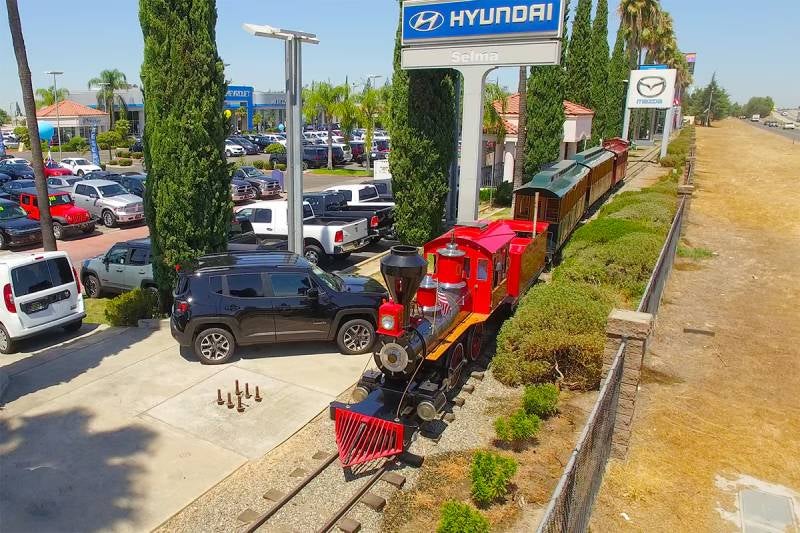 Colorful mini train displayed on tracks beside Selma Hyundai dealership, with parked cars, tall trees, and dealership signs in the background.