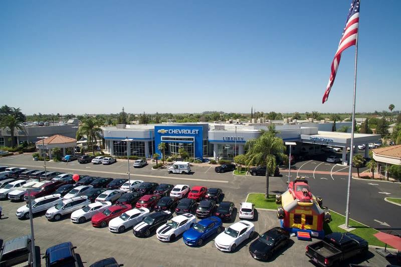 Aerial daytime view of a Chevrolet dealership with rows of parked cars, palm trees, a large American flag, and a small inflatable bounce house in the parking lot.