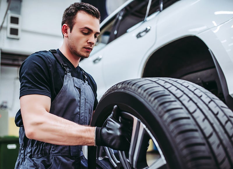 A mechanic in gray overalls and a black shirt, wearing black gloves, lifts a car wheel and tire next to a raised white SUV.